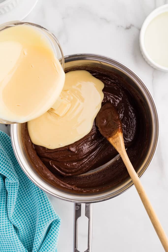 A glass bowl of sweetened condensed milk is being poured into a saucepan of melted chocolate.