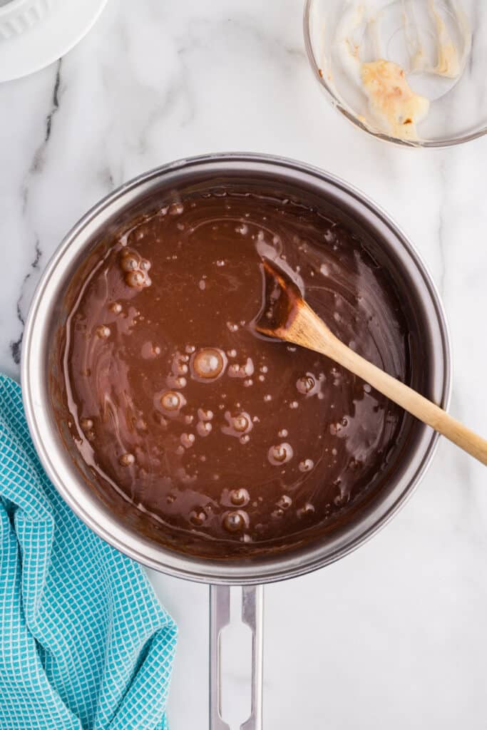 Bubbling chocolate fudge sauce in a saucepan with a wooden spoon.