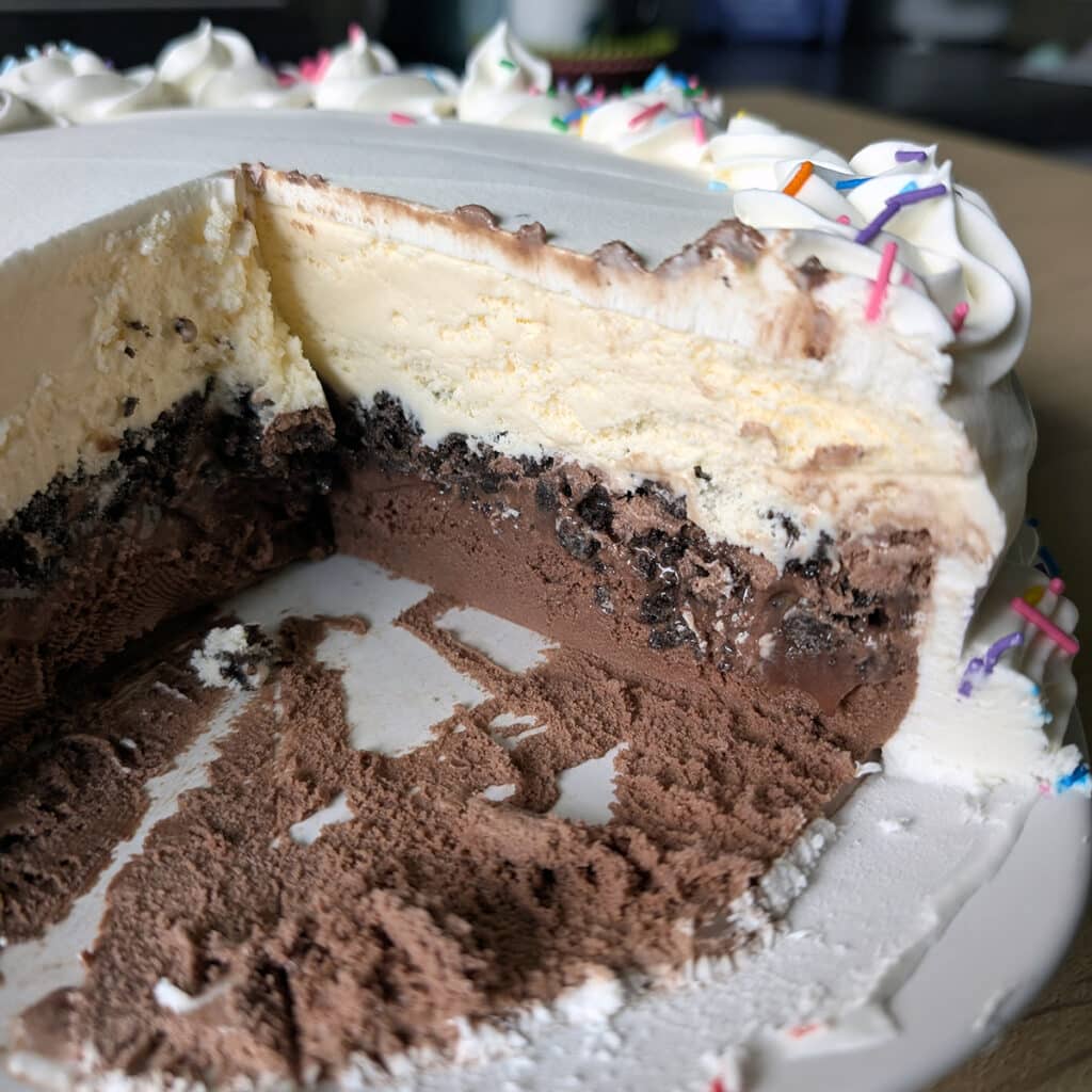 Ice cream cake with a large slice removed, showing chocolate and vanilla layers, cookie crumble, and fudge.