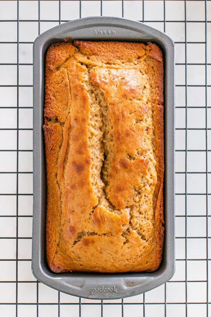 A loaf of apple cider donut bread still in the baking pan, cooling on a wire rack, with a golden-brown top and a split down the center.