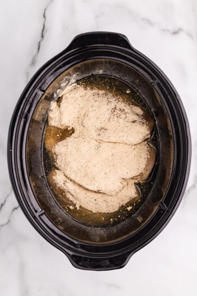 Uncooked chicken breasts topped with seasoning in a slow cooker.