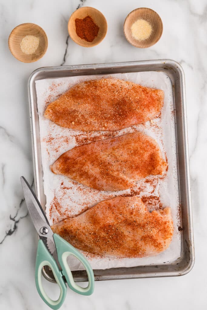Raw chicken breasts coated in spices on a parchment-lined baking tray.