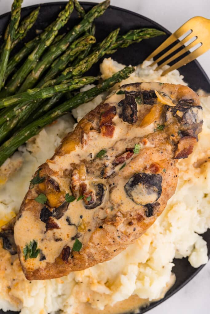 A juicy chicken breast covered in creamy sauce with mushrooms, bacon, and herbs rests on mashed potatoes beside roasted asparagus
