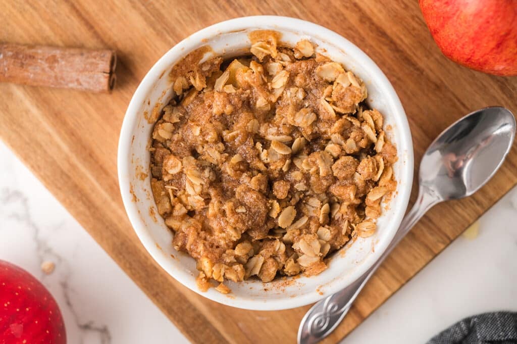 A baked apple crisp with golden oat topping in a white ramekin on a wooden board.