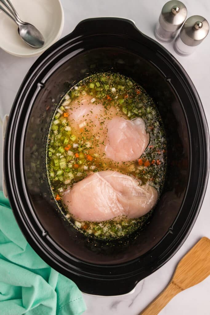 Raw chicken breasts in broth with diced vegetables inside a slow cooker.