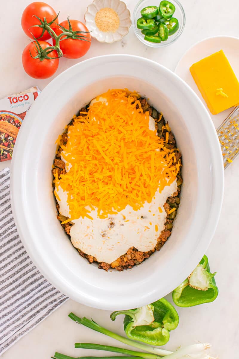 Uncooked cheesy casserole layers in a slow cooker surrounded by fresh ingredients.