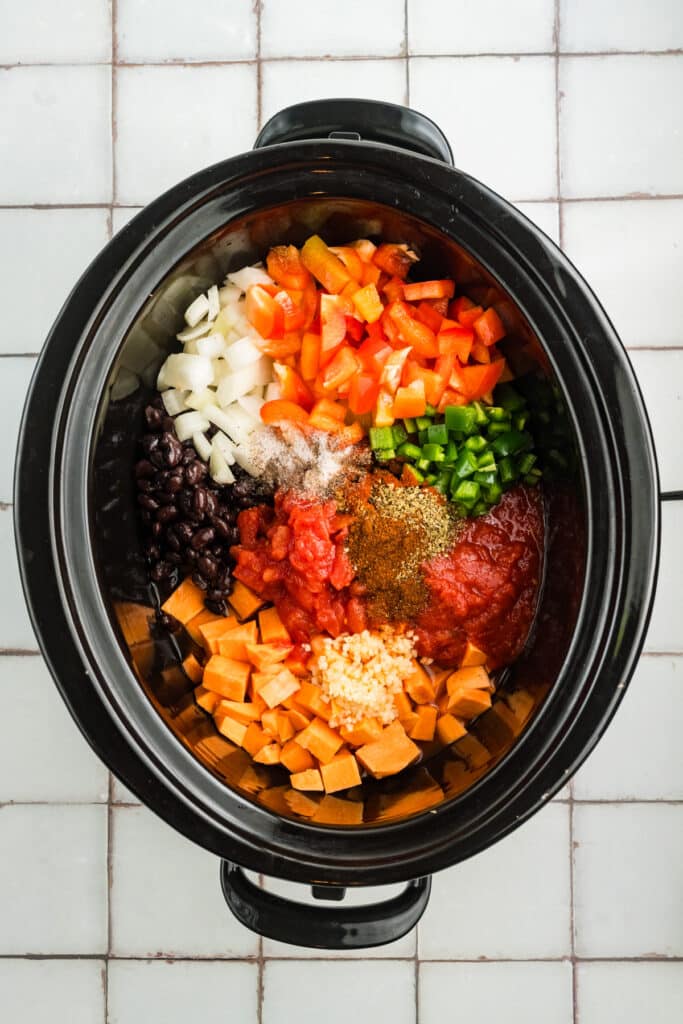 a slow cooker filled with uncooked chili ingredients including chopped sweet potatoes, bell peppers, onions, jalapeños, black beans, tomatoes, garlic, and seasonings.