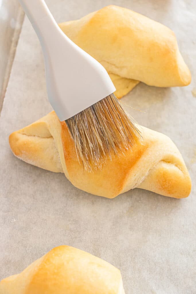 A close-up of a pastry brush spreading butter on a baked crescent roll.