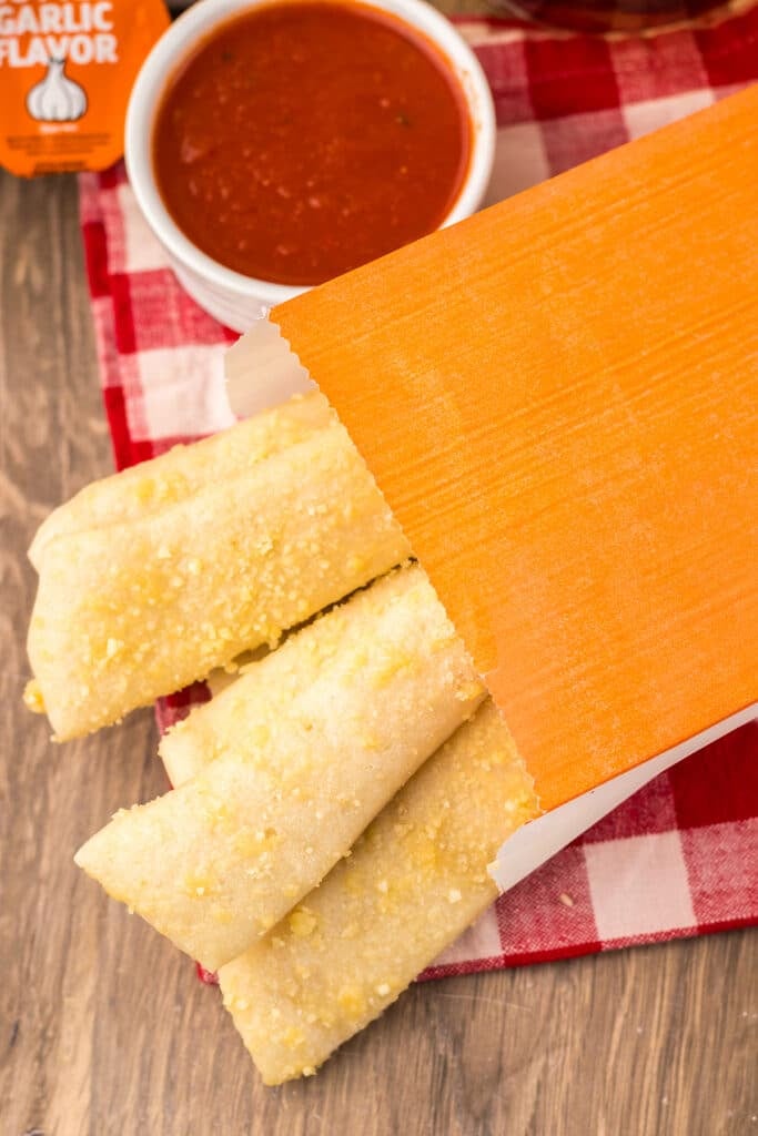 Breadsticks in an orange sleeve beside a cup of marinara sauce.