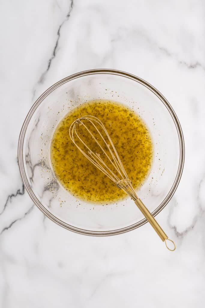 Whisk mixing oil and seasonings in a glass bowl on marble surface.