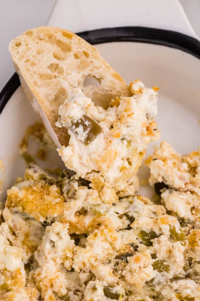 A slice of bread dipped into jalapeño popper dip inside a white baking dish.