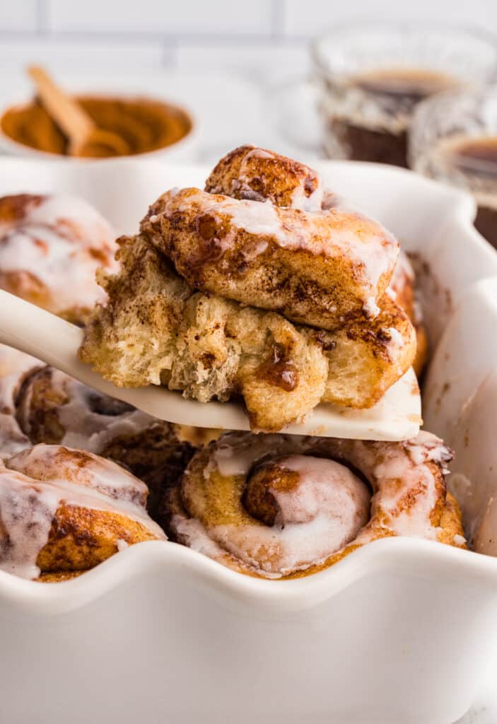 Iced cinnamon roll being lifted from a white ceramic baking dish using a spatula, with other rolls visible underneath.