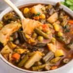 Spoon lifting chunky vegetable soup with potatoes, green beans, peas, corn, and carrots in a broth.