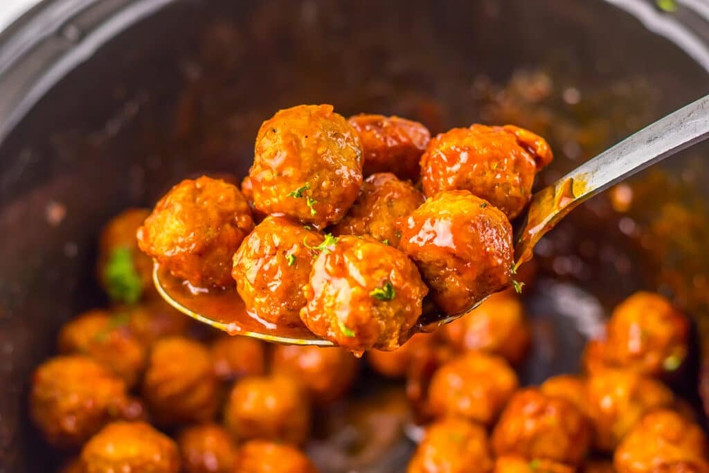 Metal spoon lifting sauced meatballs from a slow cooker.