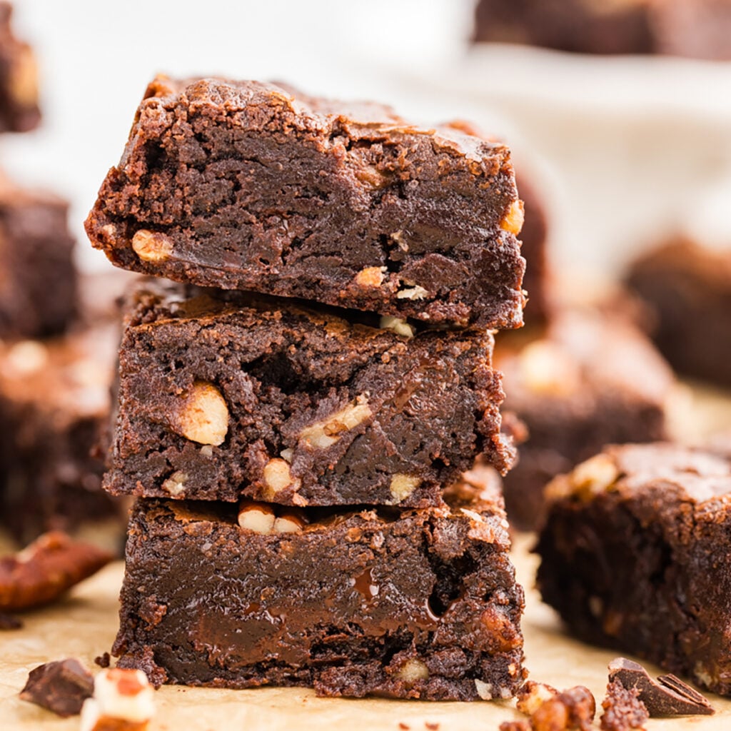 Stack of fudgy chocolate brownies with pecans and melted chocolate chunks on a wooden board.