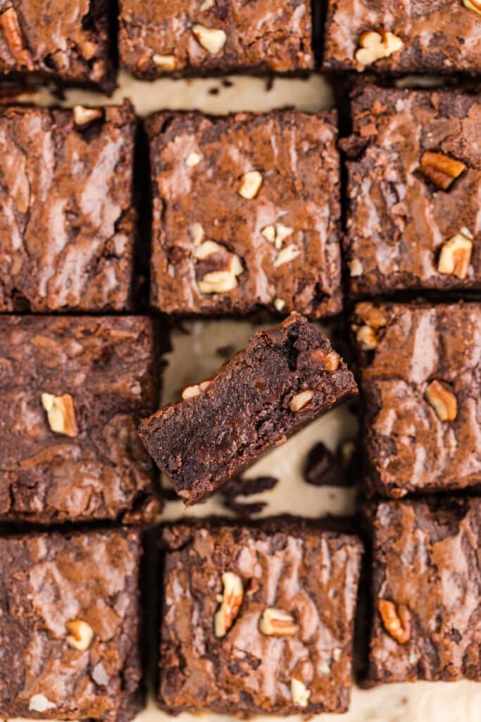 Grid of fudgy chocolate brownies topped with pecans with one brownie piece lifted to show the interior.