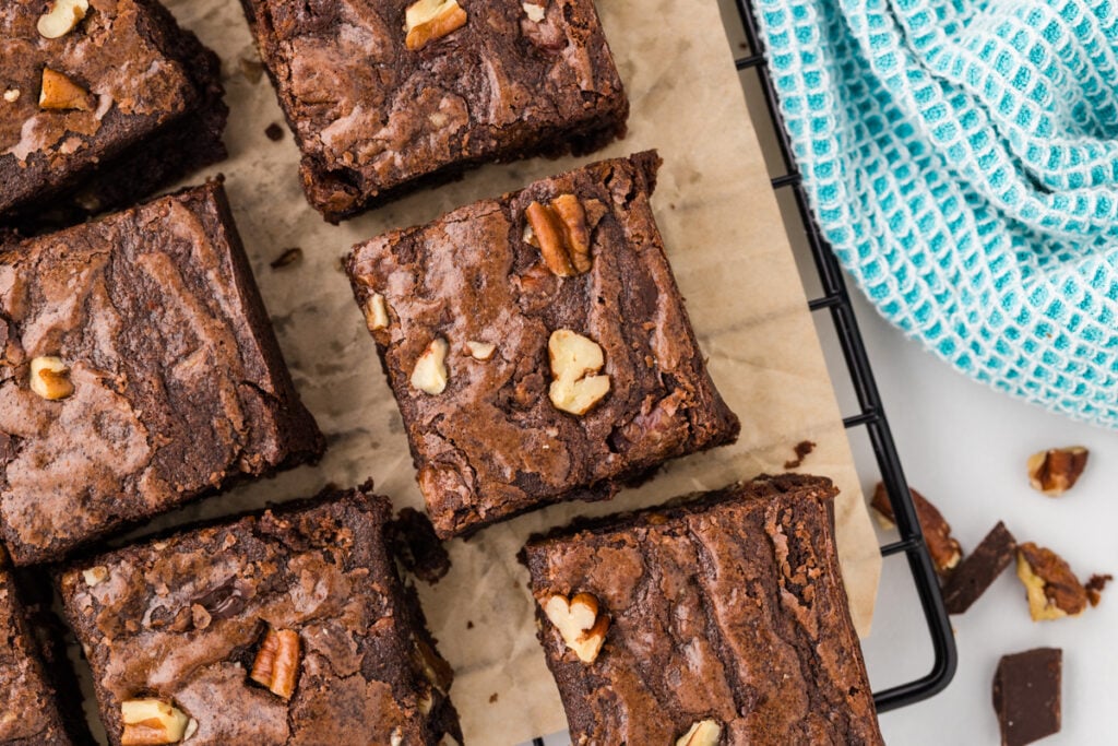 Overhead view of square chocolate brownies topped with pecan pieces arranged on parchment paper on a cooling rack.