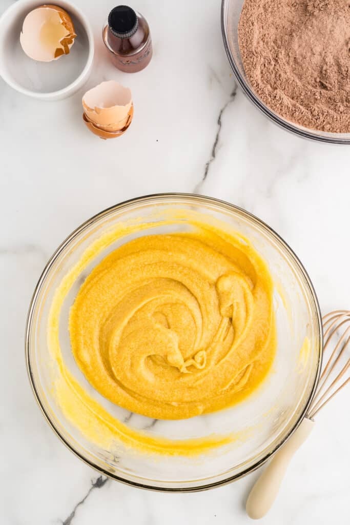 Glass bowl with yellow batter being whisked on a marble counter with eggshells, vanilla extract, and a bowl of cocoa powder nearby.