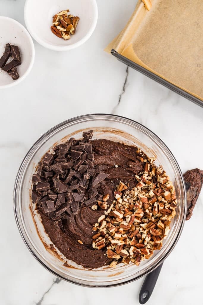 Glass bowl of chocolate brownie batter topped with chopped pecans and chocolate chunks.